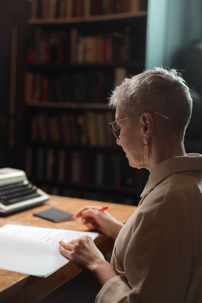 A senior woman examines a script with a red pen at a desk in a library setting.